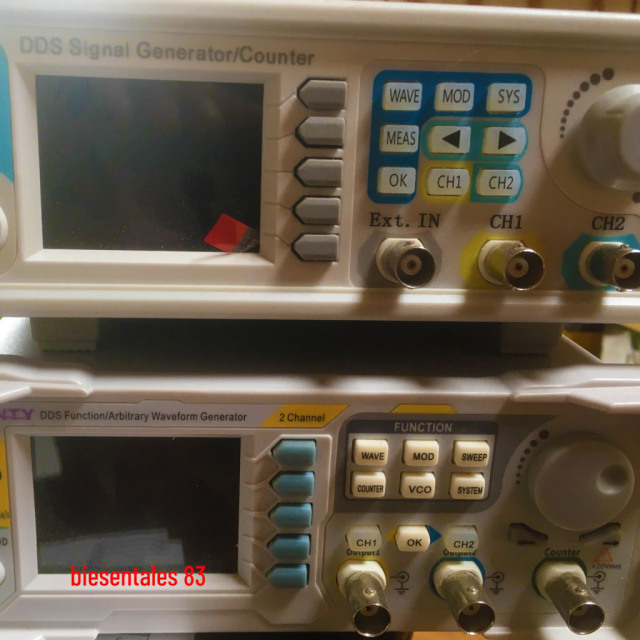 Two cheap chinese made laboratory signal generators, each with a screen, buttons. a big dial and BNC connectors in light grey plastic housing.